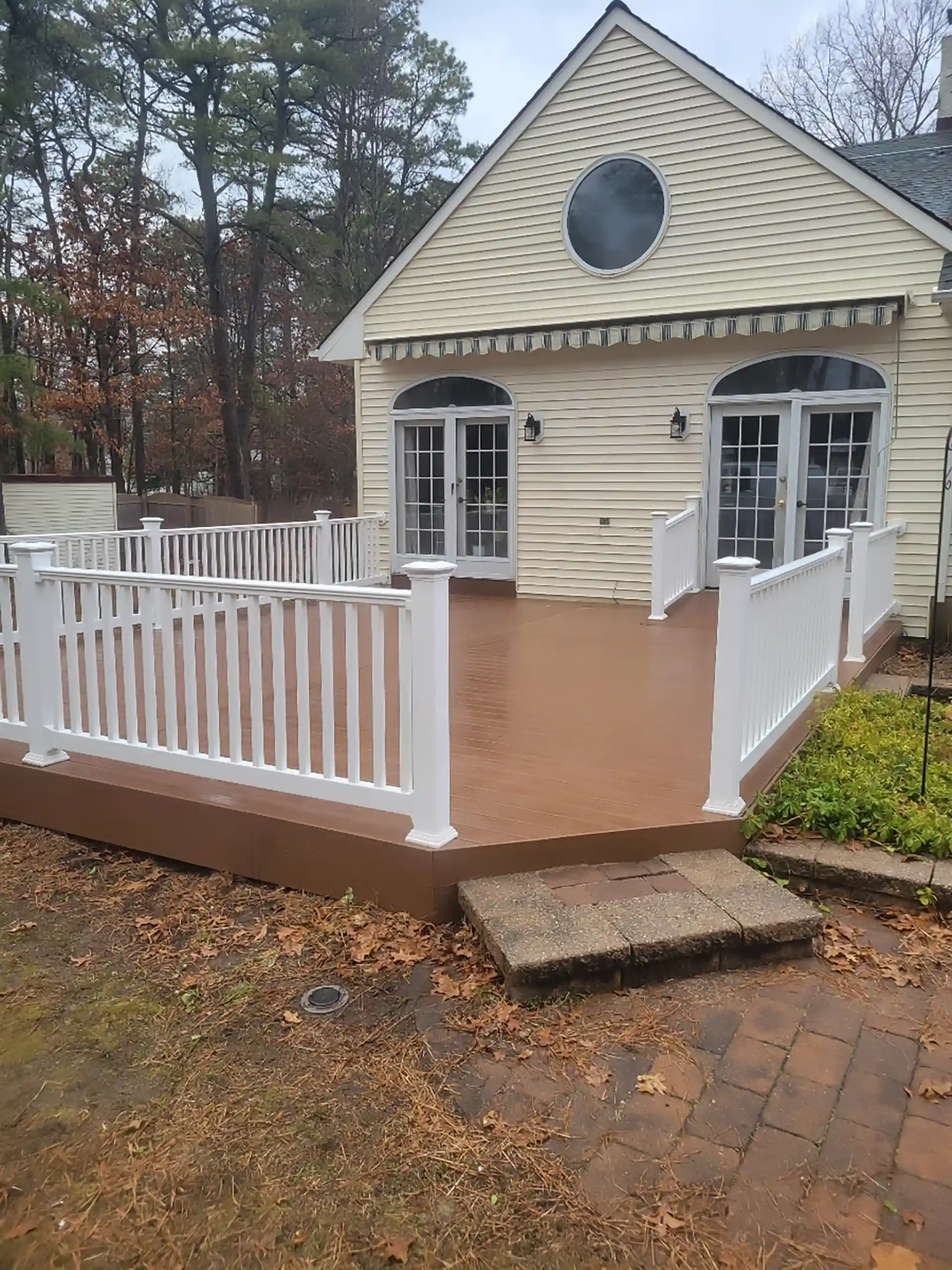 Outdoor deck with brown planks and white railings, adjacent to a yellow house with double doors and a stone step.