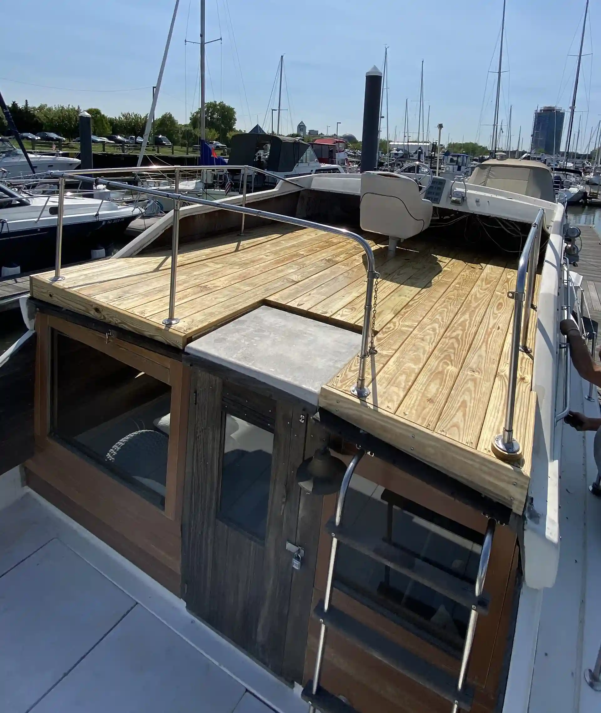 Boat rooftop deck with wood-patterned surface, metal railing, and ladder access, docked in a marina with sailboats in the background.
