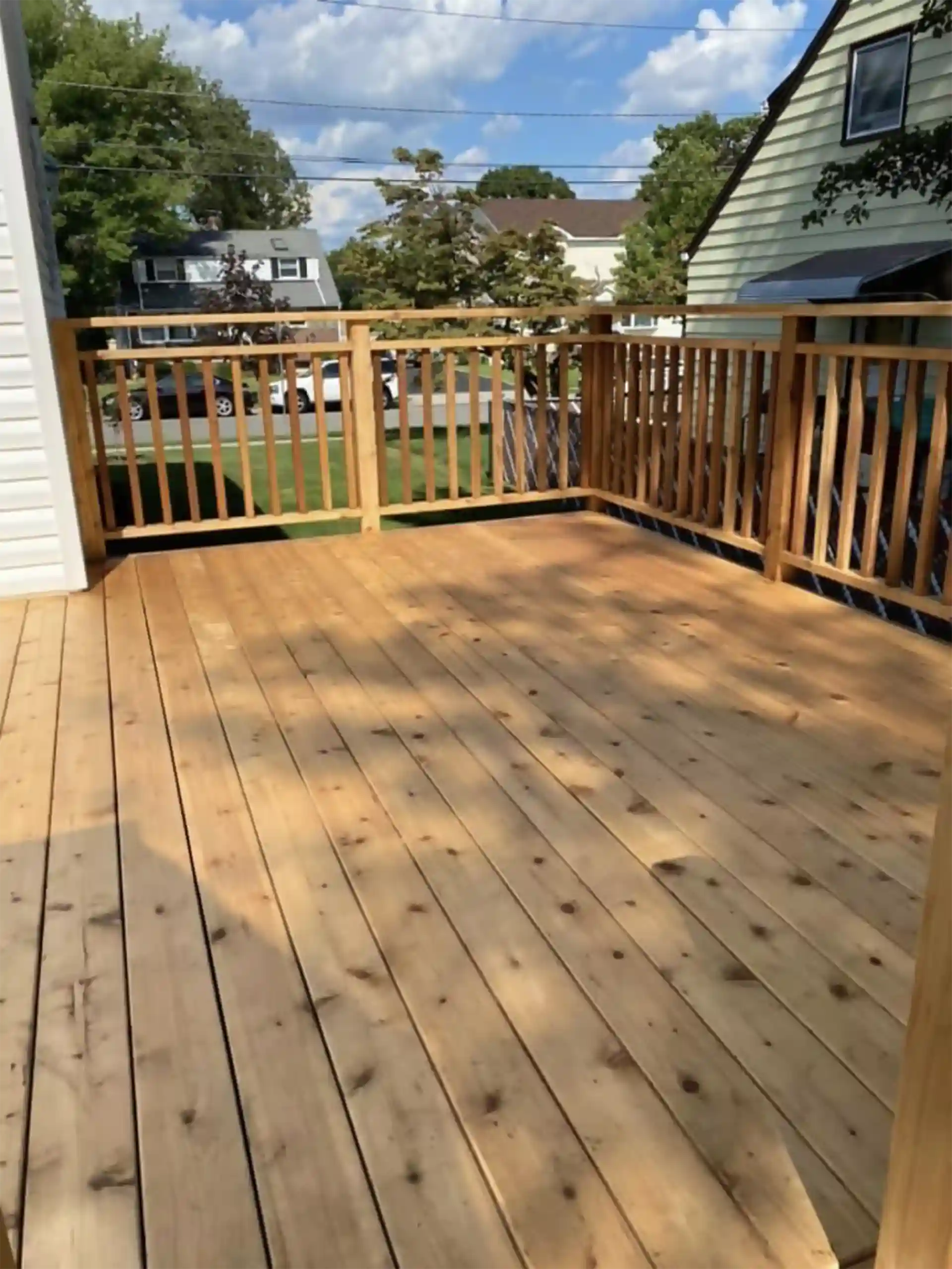Newly built natural wood deck with vertical wooden railings overlooking a green lawn and neighboring houses on a sunny day. 