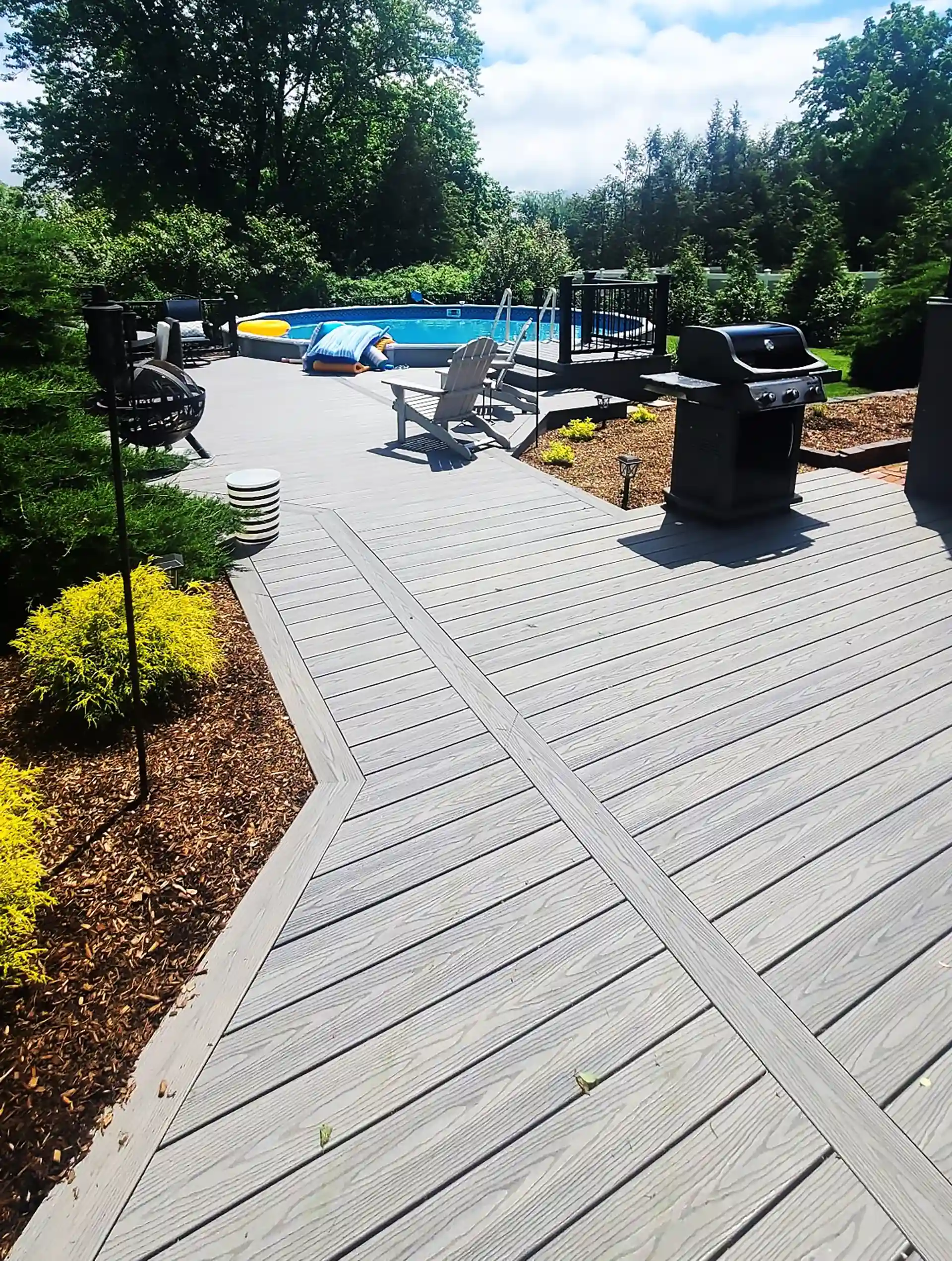 Spacious gray composite deck with built-in seating area, grill, and pool surrounded by lush greenery. 